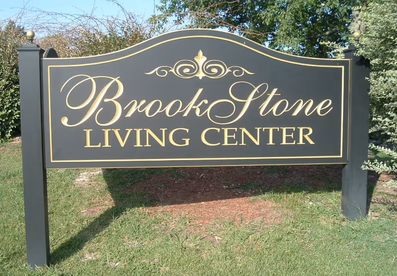 A black and gold outdoor sign for Brook Stone Living Center, mounted on two black posts with decorative finials, surrounded by grass and greenery.