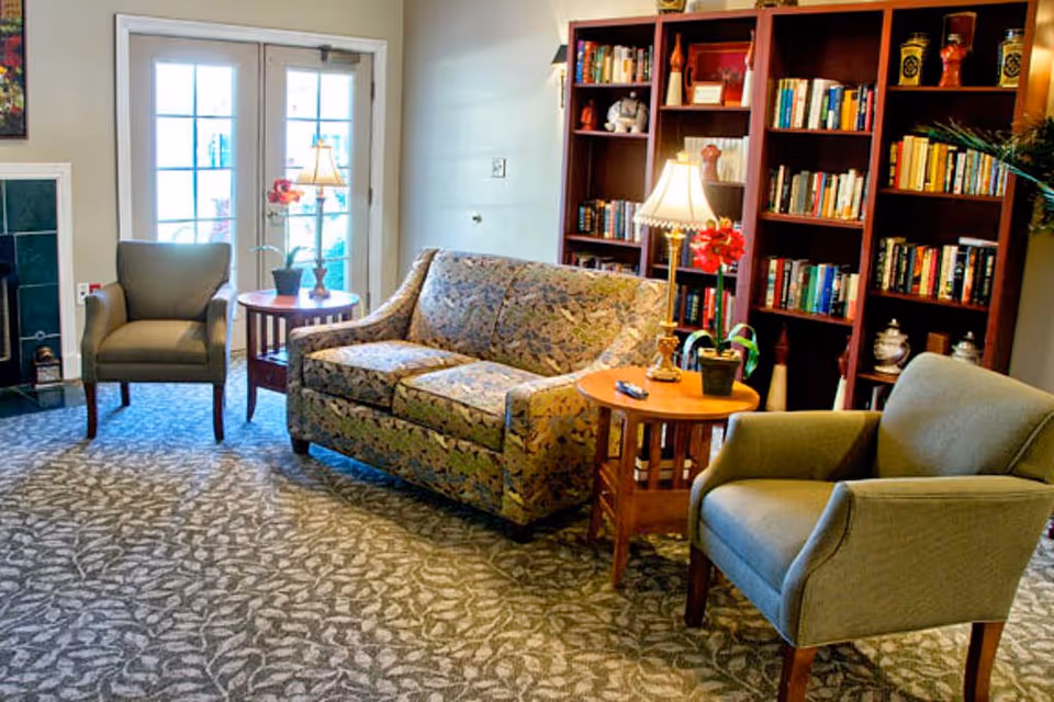 Cozy living room with a patterned sofa, two armchairs, side tables with lamps, and a bookshelf filled with books.