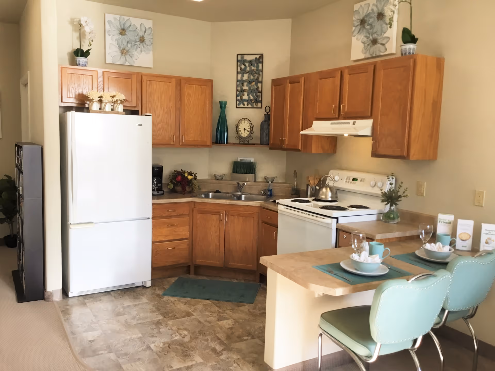 A cozy kitchen area with wooden cabinets, a white refrigerator, a white stove with a kettle on it, and a double sink. The countertop extends to form a small breakfast bar with two teal cushioned chairs. The kitchen is decorated with floral artwork, vases, and small plants. There are place settings with plates, cups, and napkins on the breakfast bar.
