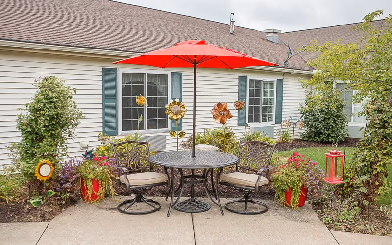 Outdoor patio area with a round metal table and four cushioned chairs under a red umbrella. The patio is surrounded by flower beds with colorful flowers, decorative metal sunflowers, and green shrubs. The background shows the exterior wall of a building with windows and green shutters.