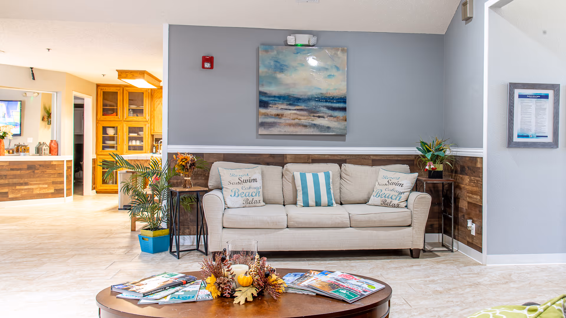 A bright and welcoming living room area in an assisted living facility featuring a beige sofa with three decorative pillows, a wooden coffee table with magazines and a fall-themed centerpiece, potted plants on either side of the sofa, a large abstract painting on the gray wall behind the sofa, and a glimpse into a kitchen area with wooden cabinets in the background.