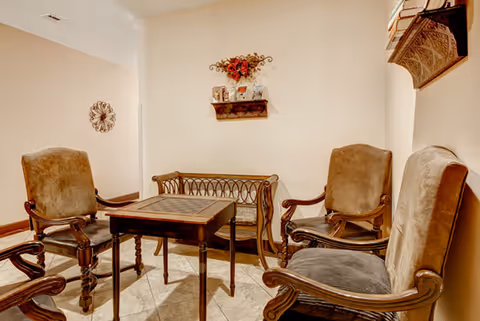 A cozy sitting area with four vintage-style wooden armchairs upholstered in brown fabric arranged around a small square table with a checkerboard pattern on top. Behind the table is a decorative wooden bench with a metal backrest. The walls are beige and decorated with a small shelf holding framed photos and a floral arrangement, and a round wall ornament is visible in the background.