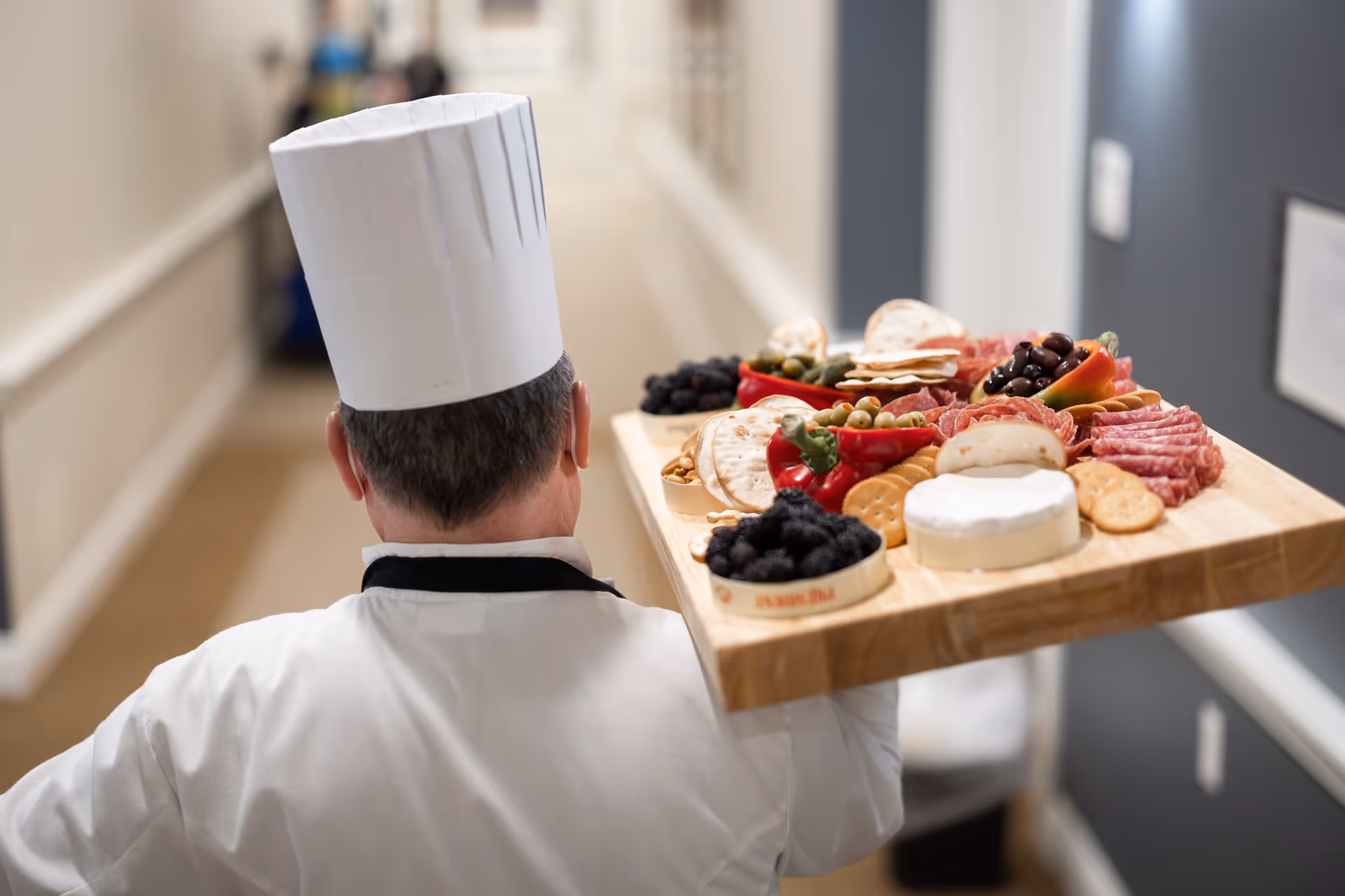 A chef wearing a white uniform and tall white hat is carrying a wooden tray filled with an assortment of foods including cheese, crackers, olives, sliced meats, and berries down a hallway.