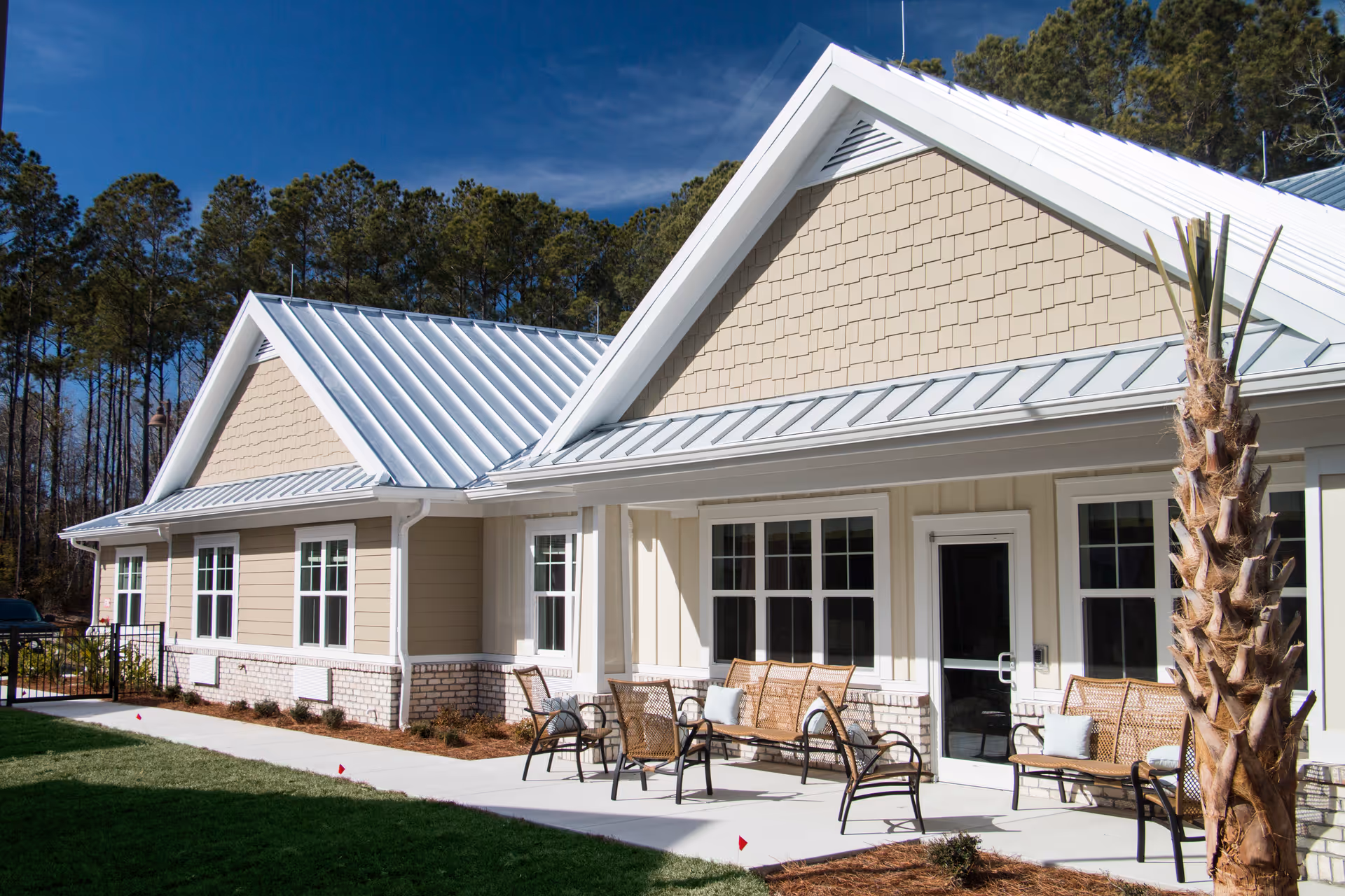 Exterior view of a senior living facility building with beige siding, white trim, and a metal roof. There are several windows and a glass door leading to a patio area with wicker chairs and benches with cushions. A palm tree is visible on the right side, and tall pine trees are in the background under a clear blue sky.