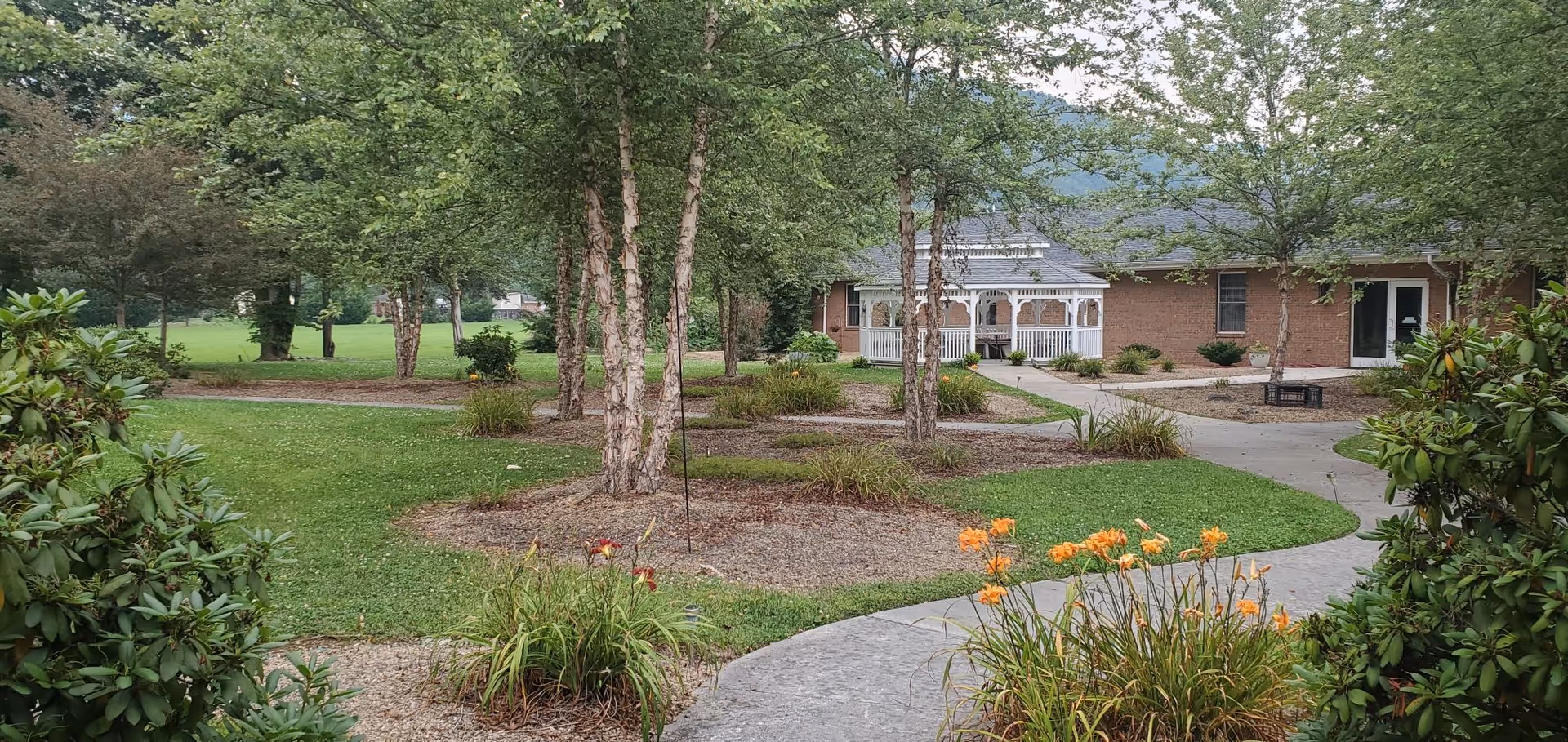 A landscaped outdoor area with green grass, trees, and flower beds featuring orange and red flowers. A paved walkway curves through the garden leading to a brick building with a white gazebo-like porch. The scene is peaceful with a backdrop of hills and cloudy sky.
