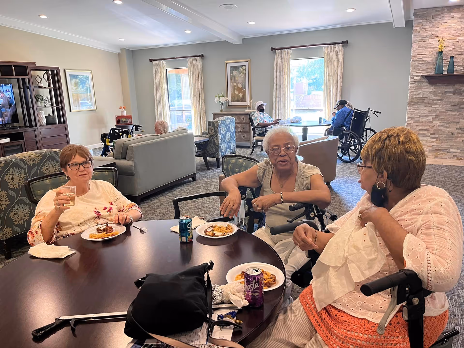 Three elderly women sitting around a round table in a common area, eating and drinking. The room has large windows with curtains, comfortable chairs, and a stone fireplace. Other residents are visible in the background, some in wheelchairs, enjoying the space.