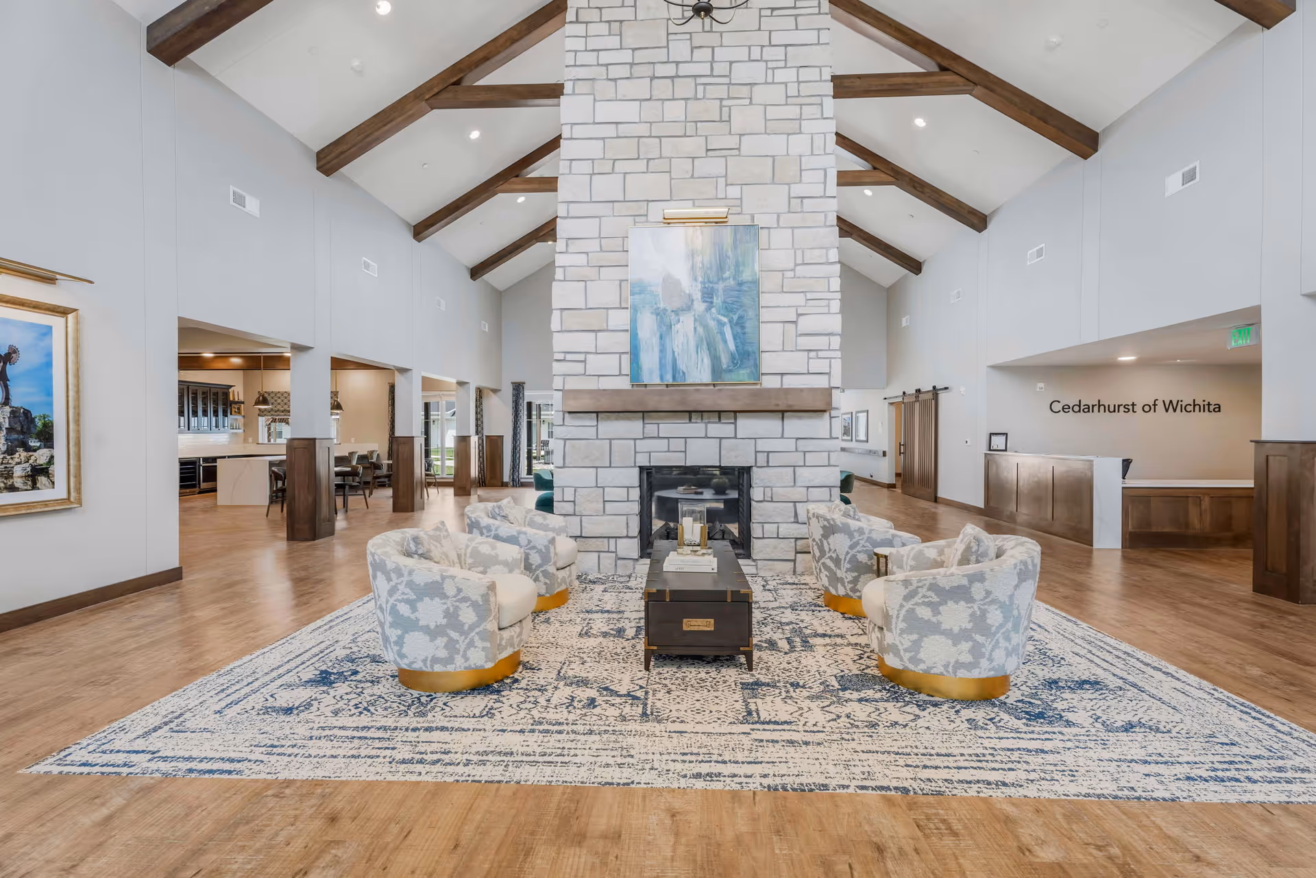 Spacious senior living facility common area with high vaulted ceilings and exposed wooden beams. Central stone fireplace with a painting above it, surrounded by four patterned armchairs on a large blue and white area rug. Wooden floors and light-colored walls with open views into adjacent dining and reception areas.