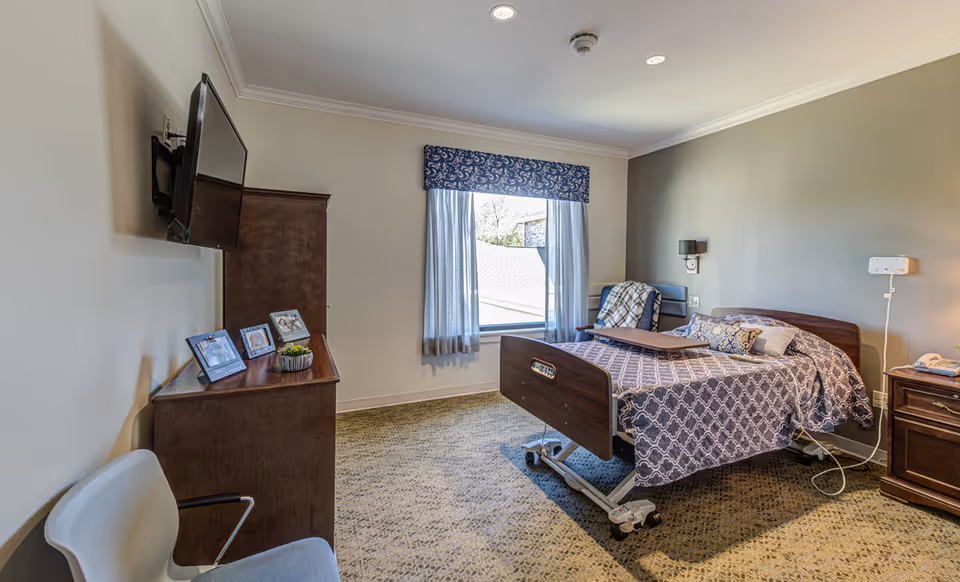 A well-lit bedroom in a senior living facility featuring a hospital-style bed with patterned bedding, a wooden nightstand with a telephone, a wooden dresser with framed photos and a small plant, a wall-mounted flat-screen TV, a window with sheer curtains and a decorative valance, and a chair with a blanket draped over it.