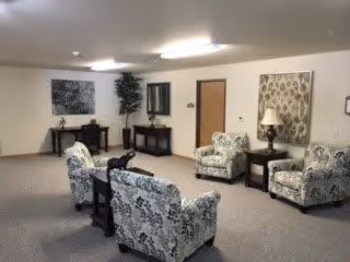 A common area in an assisted living facility featuring patterned armchairs arranged around a small table, a side table with a lamp, a large decorative wall hanging, and a console table with a plant and framed artwork on the wall.