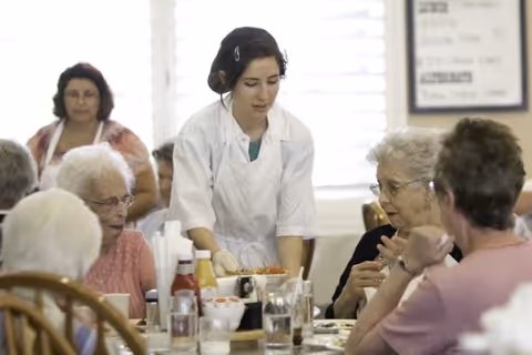 A staff member serves food to elderly residents seated around a communal dining table.