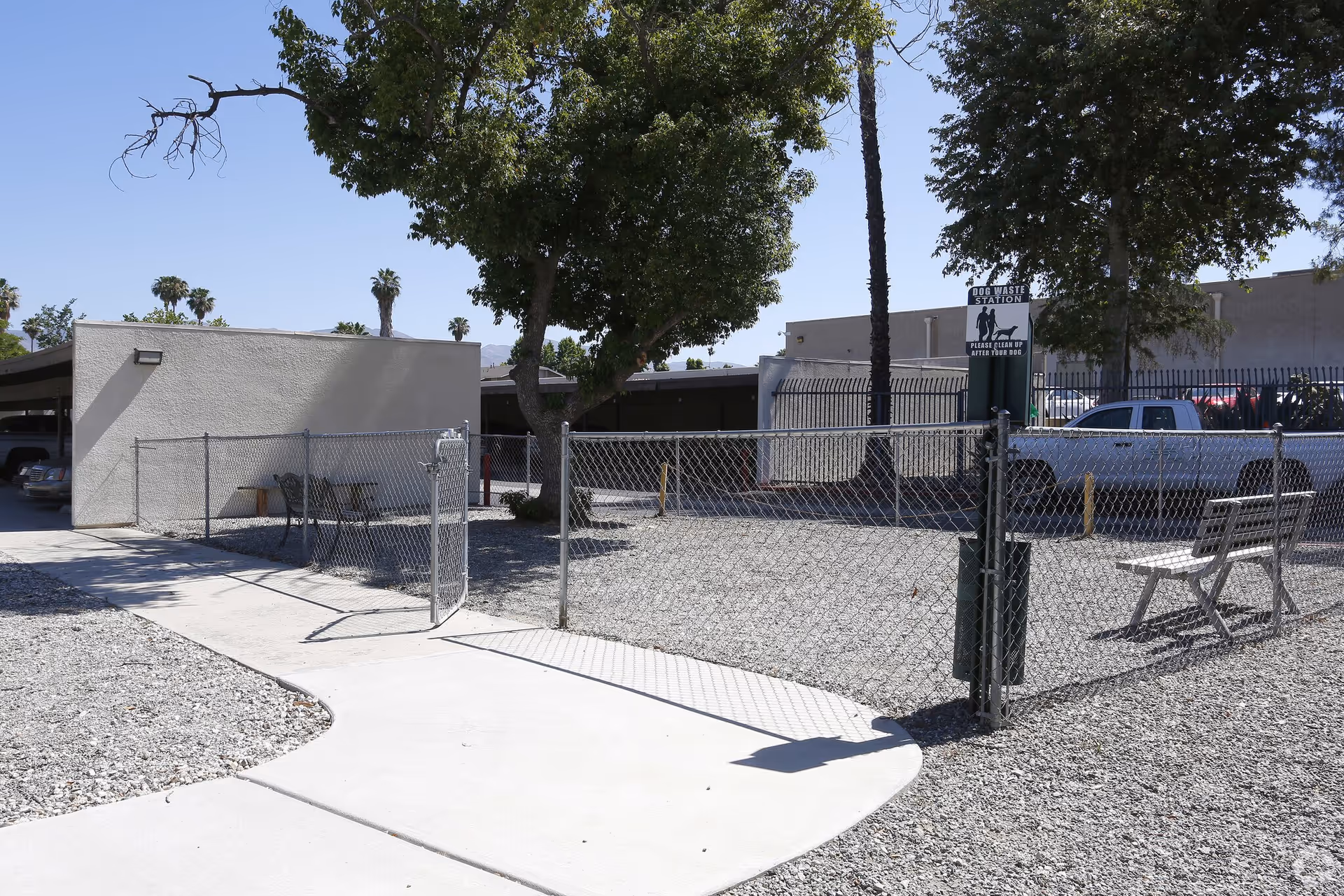 Outdoor area with a fenced dog waste station, a bench, and a few trees. There is a sidewalk leading to a gate in the chain-link fence. In the background, there are parked cars and a building under a clear blue sky.