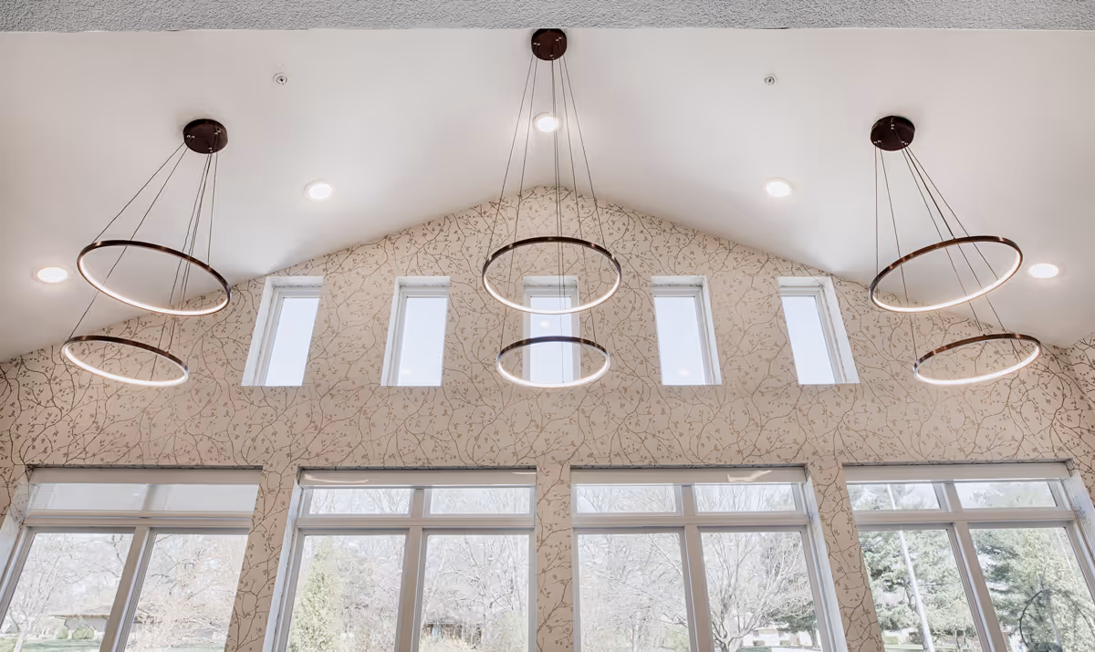Interior view of a room with a high vaulted ceiling featuring six narrow vertical windows near the top and large windows below. The walls have a light-colored wallpaper with a subtle vine pattern. Five modern circular pendant lights hang from the ceiling, emitting a soft glow.
