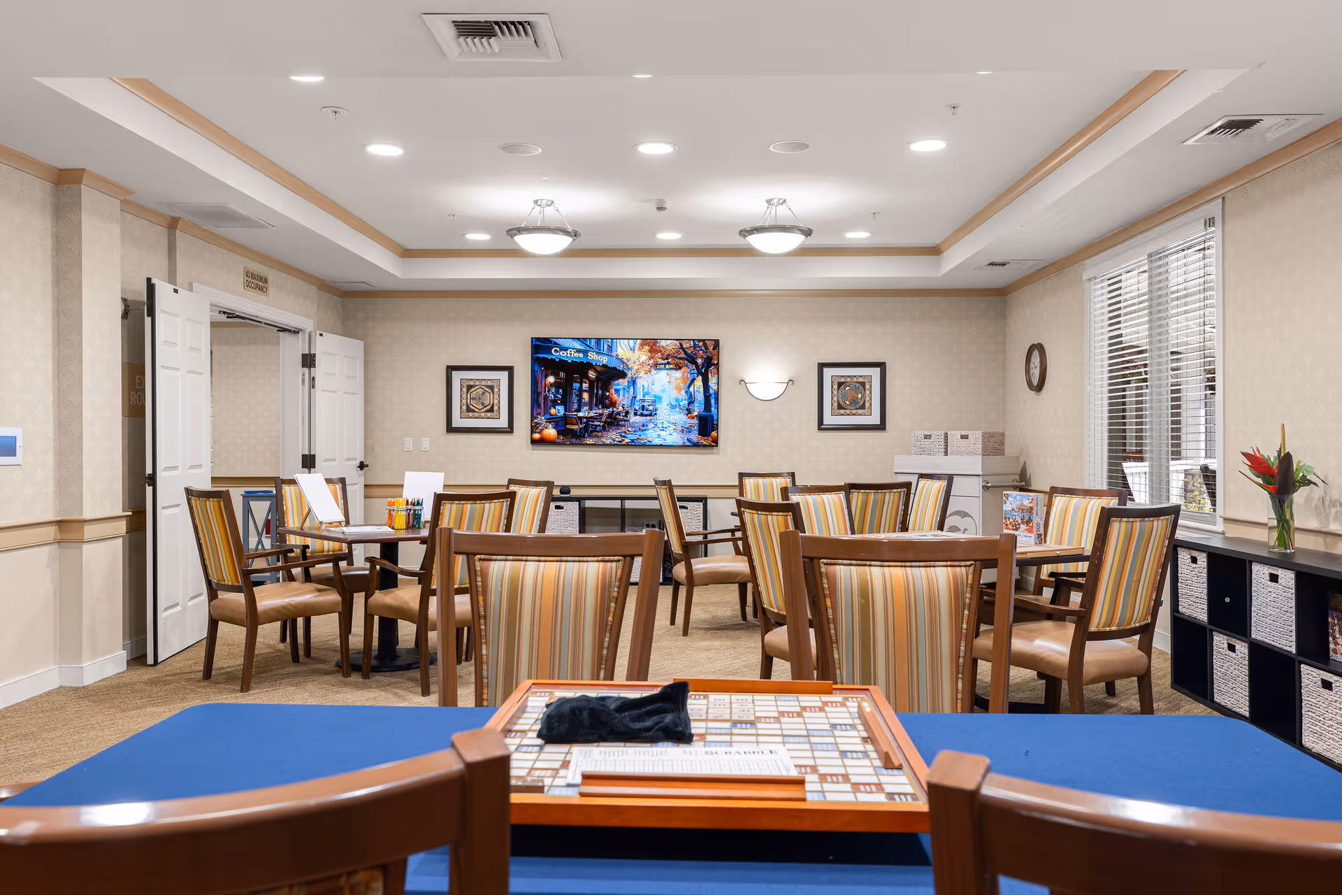 Communal dining/activity room with multiple round tables and striped chairs, a wall-mounted TV, and a Scrabble board on a front table.