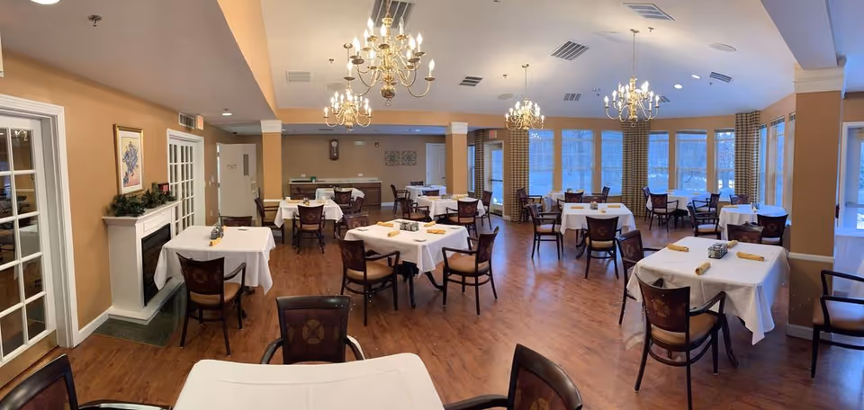 A spacious dining room with multiple tables covered in white tablecloths, each set with rolled yellow napkins and small centerpieces. The room features wooden floors, several chandeliers hanging from the ceiling, large windows with checkered curtains, and a fireplace on the left wall. The walls are painted a warm beige color.