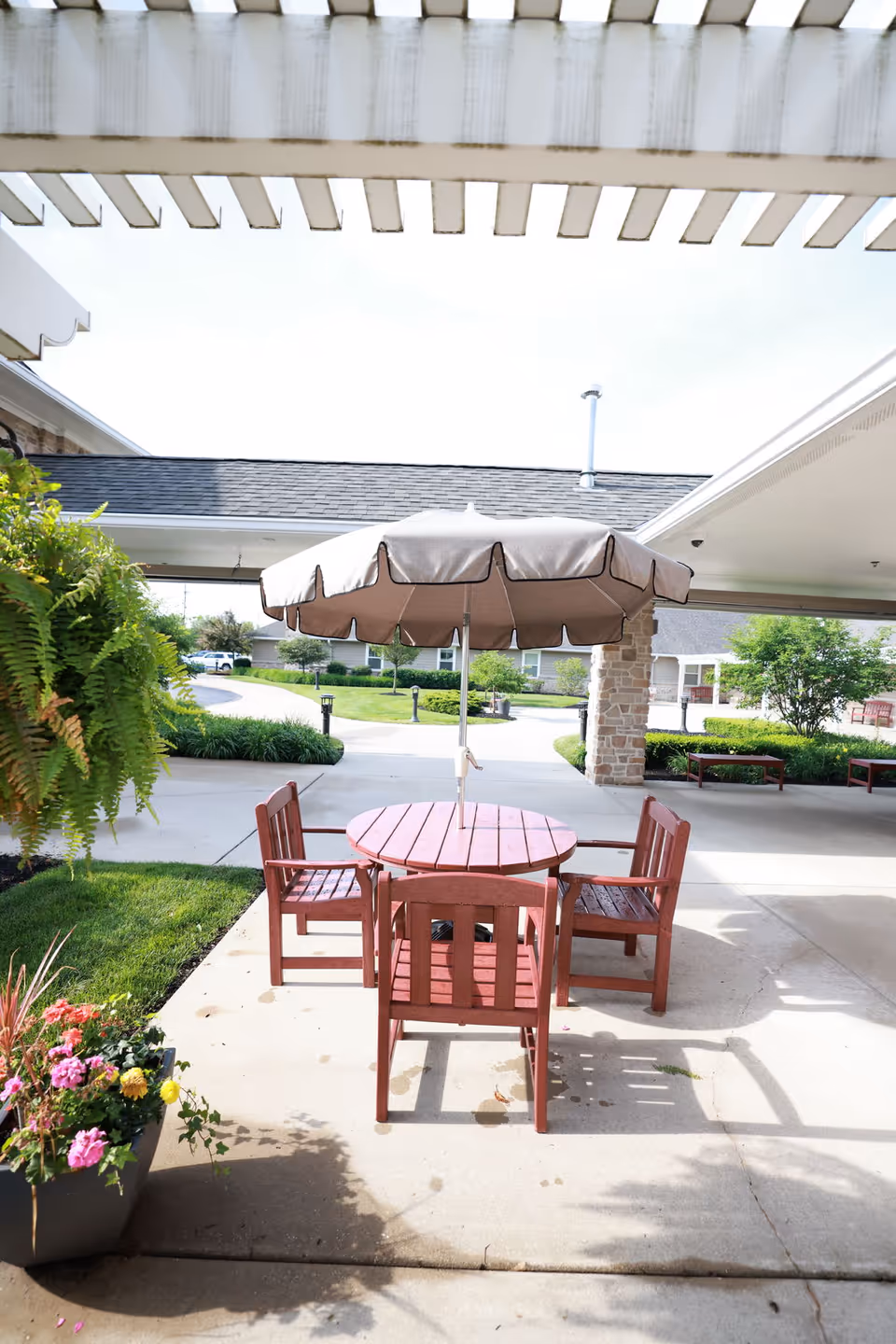 Outdoor patio with a round wooden table, four chairs and a center umbrella under a pergola near the facility entrance.