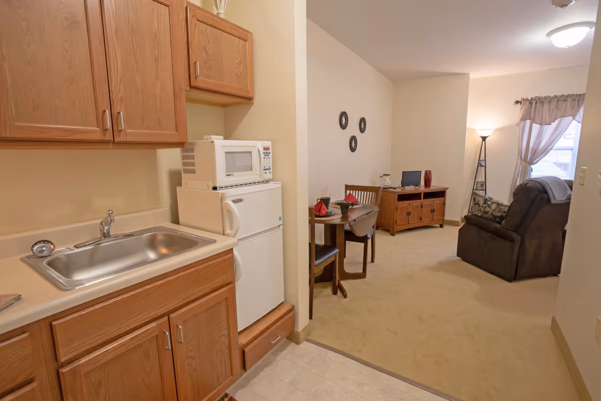 A small kitchenette with wooden cabinets, a stainless steel sink, a white microwave on top of a white mini refrigerator. Adjacent to the kitchenette is a living area with a small round dining table set for two, a wooden TV stand with a small television, a floor lamp, and a recliner chair near a window with curtains.
