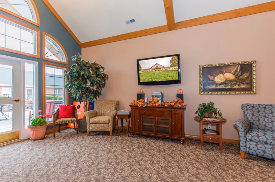 A cozy living room area with patterned carpet, a large potted plant, two armchairs, a wooden side table, and a wooden cabinet decorated with autumn leaves and small pumpkins. A flat-screen TV is mounted on the wall above the cabinet, displaying an image of a building with a green lawn. There is a framed floral painting on the wall and large windows and a door letting in natural light from outside.