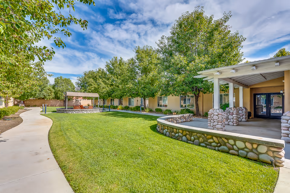 Landscaped courtyard with a winding walkway, lawn, trees, and a covered seating area outside the senior living building.