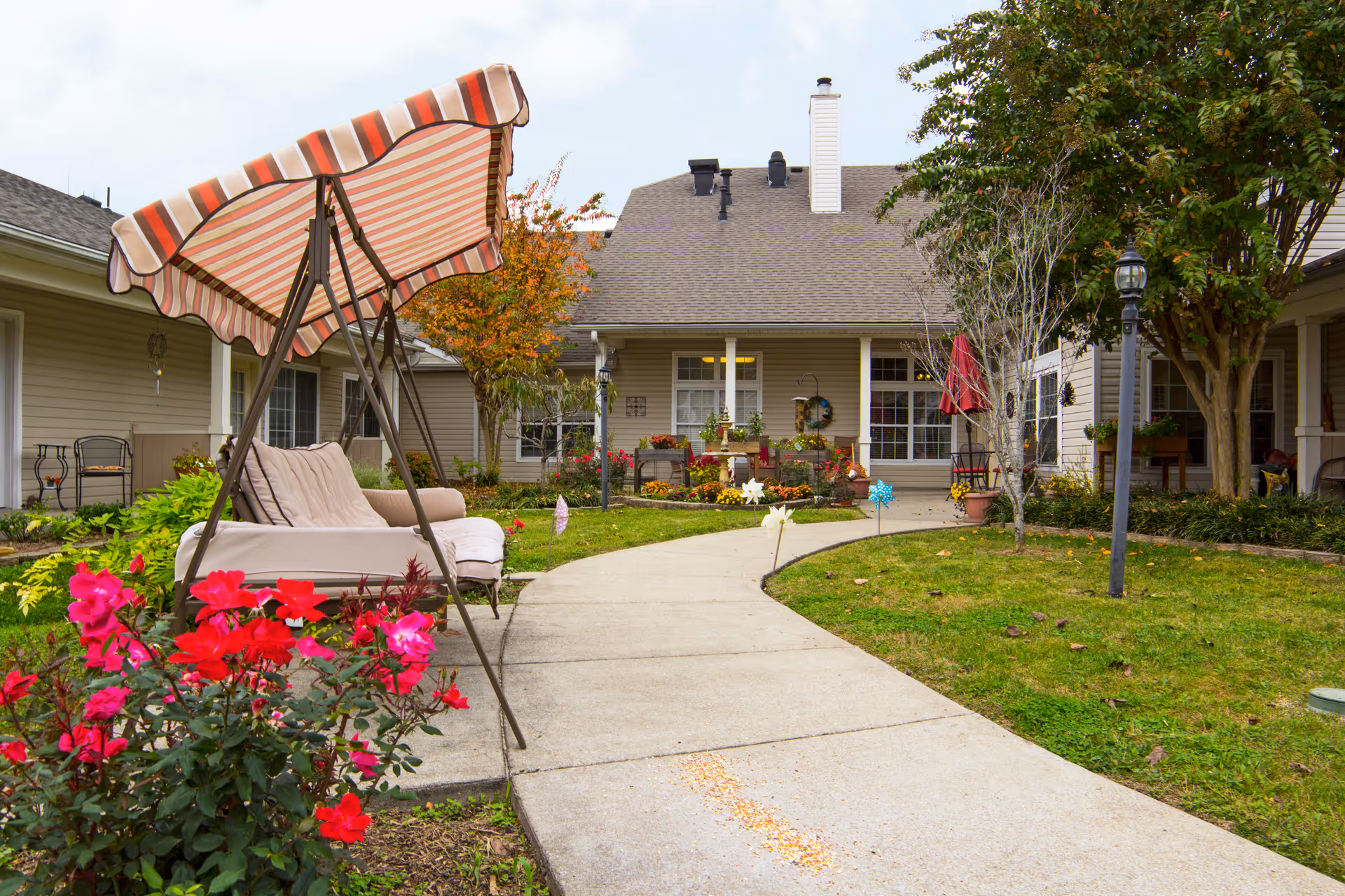 A landscaped courtyard with a striped canopy swing, paved walkway, flowering bushes, and the entrance to a single-story senior living building.
