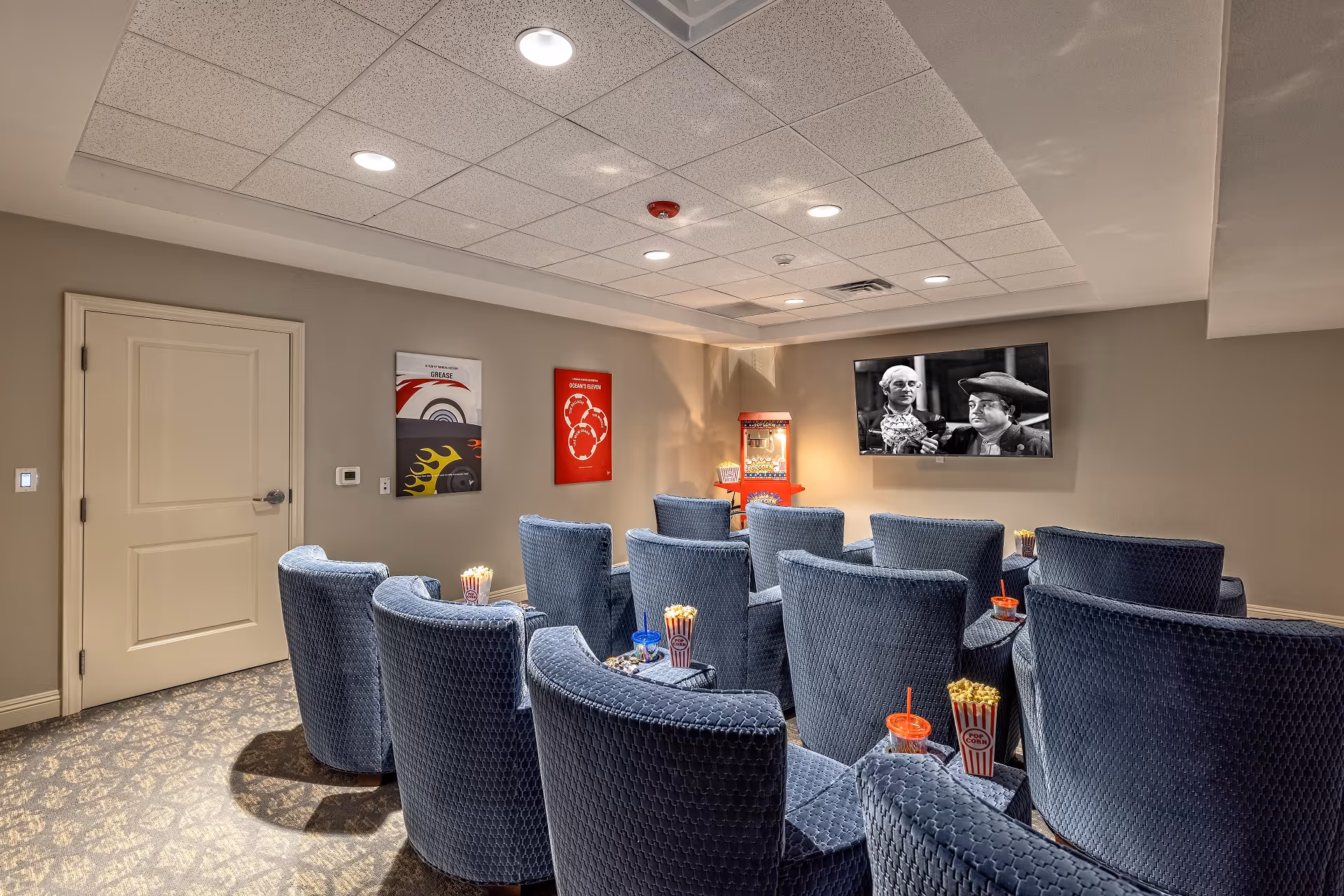 A small theater room with multiple rows of blue upholstered chairs facing a wall-mounted TV screen showing a black and white movie. There is a popcorn machine in the corner and several popcorn containers and drinks placed on the armrests of the chairs. The room has beige walls, a patterned carpet, and recessed ceiling lights.