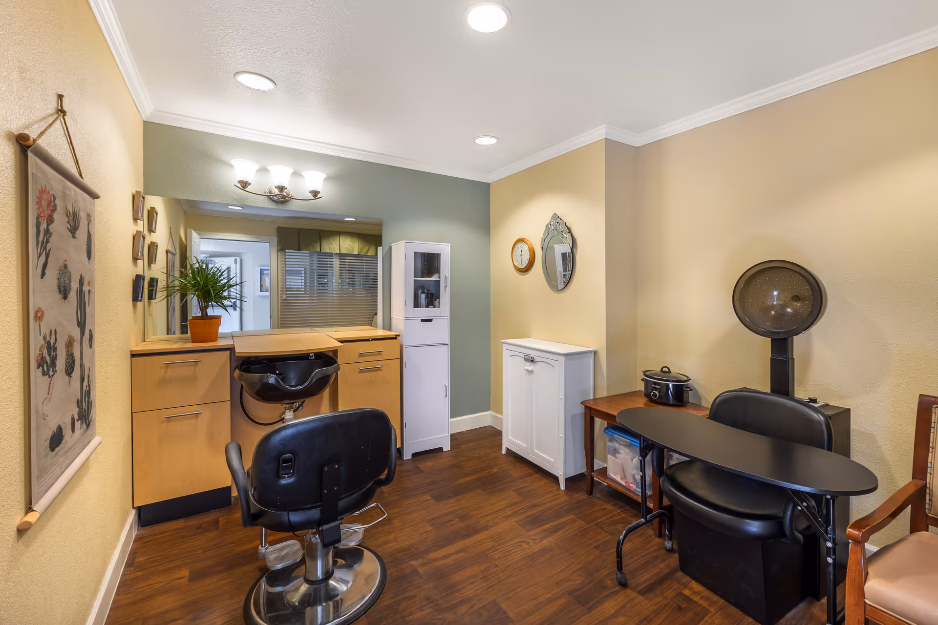Small salon-style interior with styling chairs, a shampoo sink, a hair dryer hood, cabinetry and a mirror.