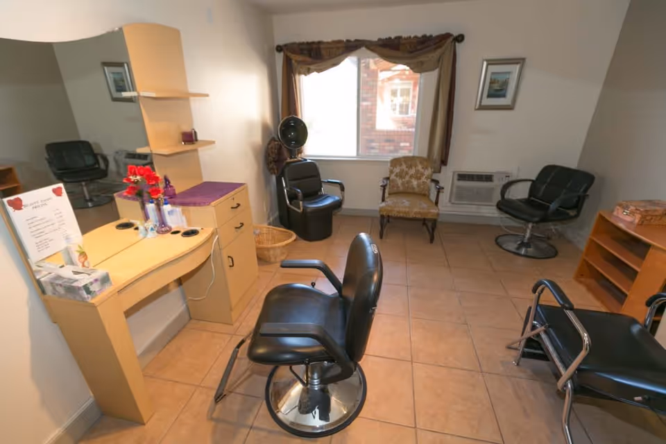 Interior room with salon-style chairs, a vanity with a large mirror, a window with curtains, and a few other chairs arranged around the room. The floor is tiled and there is an air conditioning unit below the window.