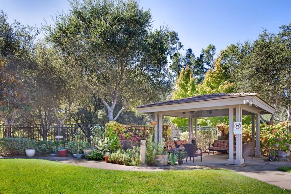 Outdoor garden area with a covered seating pavilion containing cushioned chairs and a sofa. The area is surrounded by green grass, various plants, and trees under a clear blue sky.
