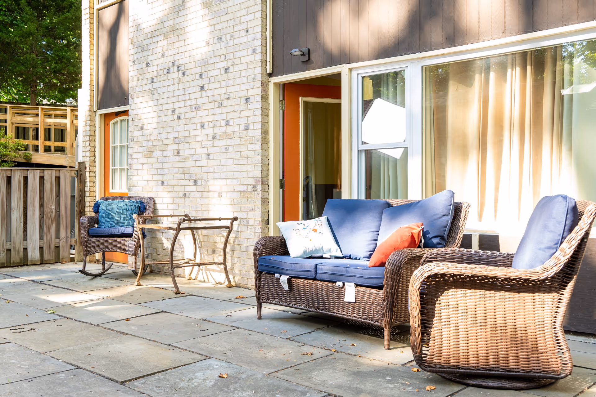 Outdoor patio area with wicker furniture including a loveseat with blue cushions and pillows, a matching armchair, and a rocking chair with a blue cushion. The patio is paved with stone tiles and is adjacent to a building with a brick and wood exterior. There is a glass door and window with beige curtains.