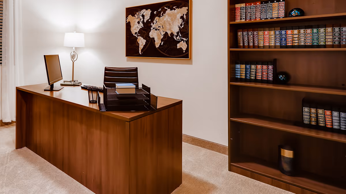 A tidy wooden office with a large desk, leather chair, lamp, computer monitor, world map on the wall, and a bookshelf filled with supplies.