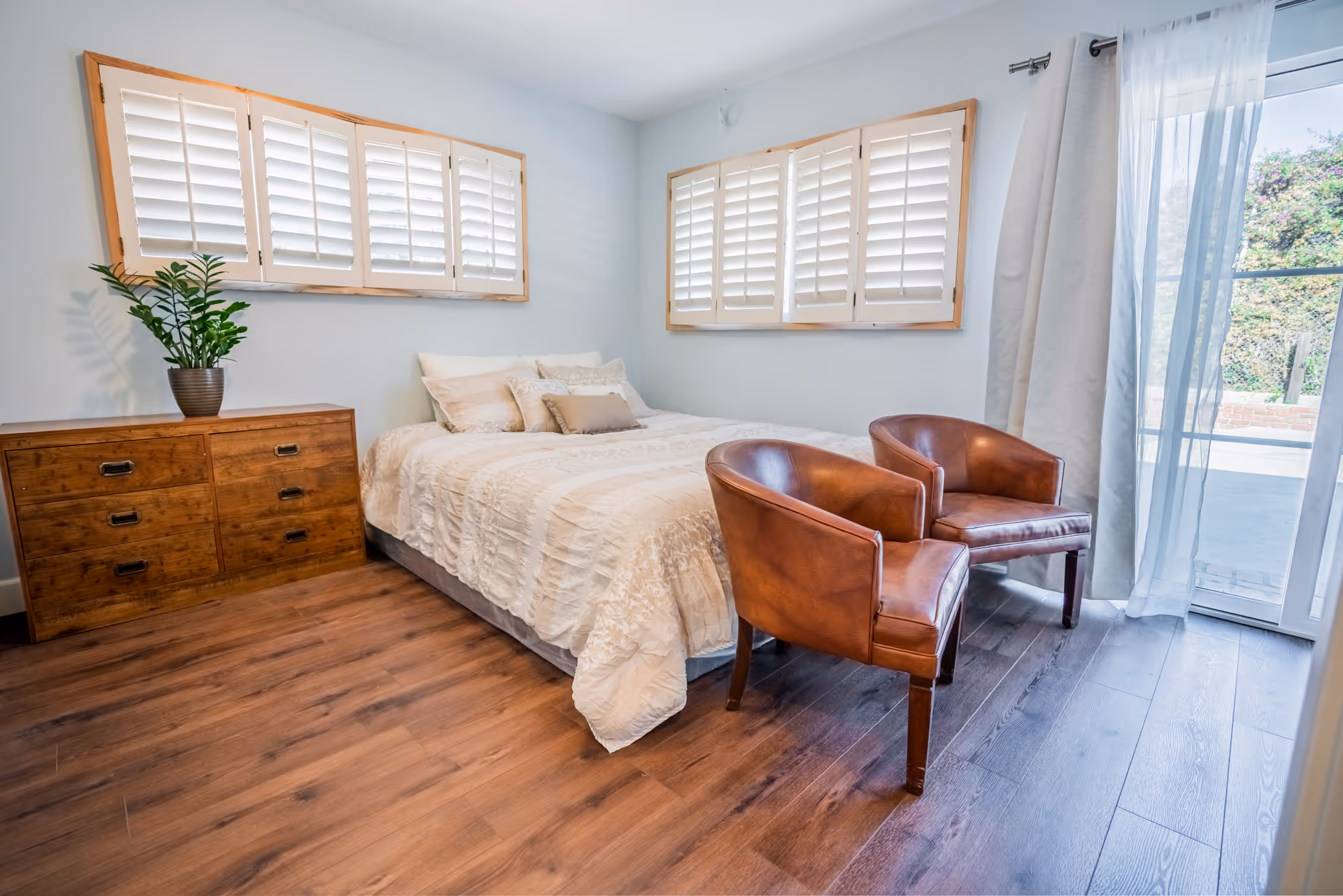 A bright bedroom with a neatly made bed featuring white bedding and multiple pillows. There are two brown leather chairs placed at the foot of the bed. A wooden dresser with a potted plant on top is positioned against the wall. The room has wooden flooring, two windows with white shutters, and a glass door with sheer curtains leading outside.