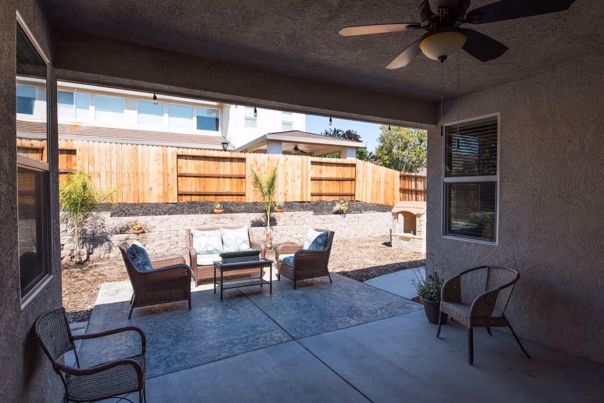 Covered outdoor patio area with ceiling fan, featuring wicker chairs and a small table. Beyond the patio is a fenced backyard with a retaining wall, small plants, and a neighboring building visible in the background.