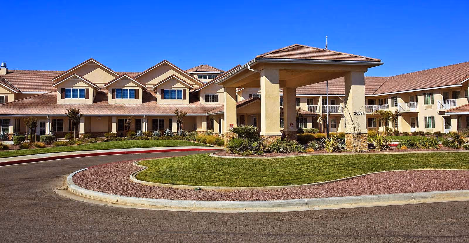 Exterior view of a senior living facility with beige walls and a red-tiled roof under a clear blue sky. The building has multiple windows and a covered entrance with landscaping and a curved driveway in front.