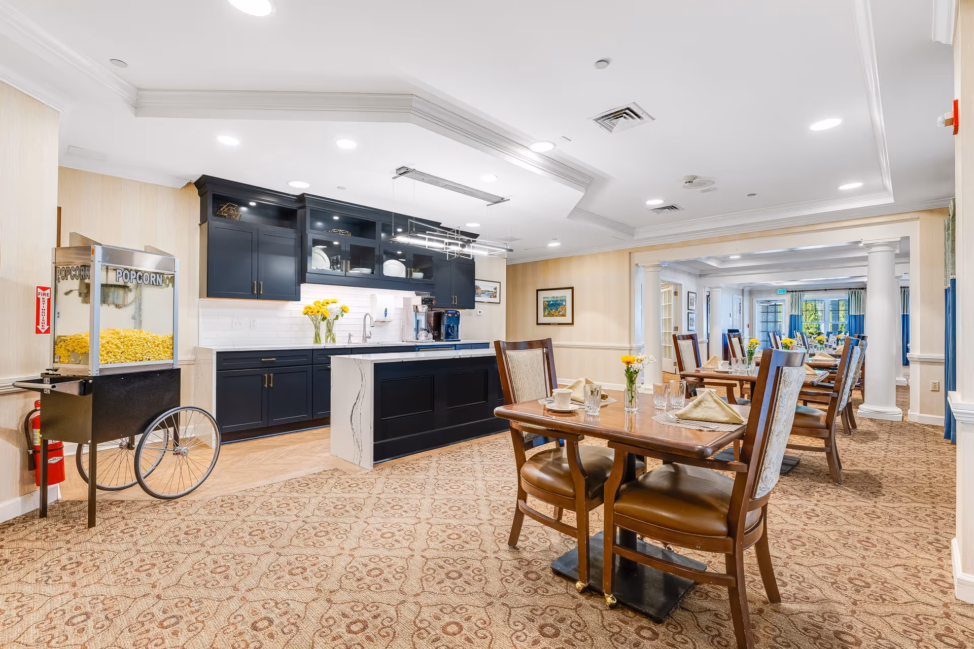 A bright dining area in a senior living facility featuring wooden tables set with glasses, napkins, and small flower vases. To the left, there is a popcorn machine and a modern kitchen area with dark blue cabinets, a white countertop island, and a coffee machine. The room has patterned carpet flooring and recessed ceiling lights.