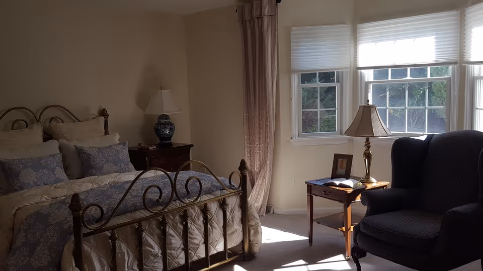 A cozy bedroom with a metal-framed bed adorned with multiple pillows and a patterned comforter. Next to the bed is a wooden nightstand with a decorative lamp. Sunlight streams through three large windows with white blinds, illuminating a wooden side table with a lamp, an open book, and a framed photo. A dark upholstered armchair is positioned near the side table.