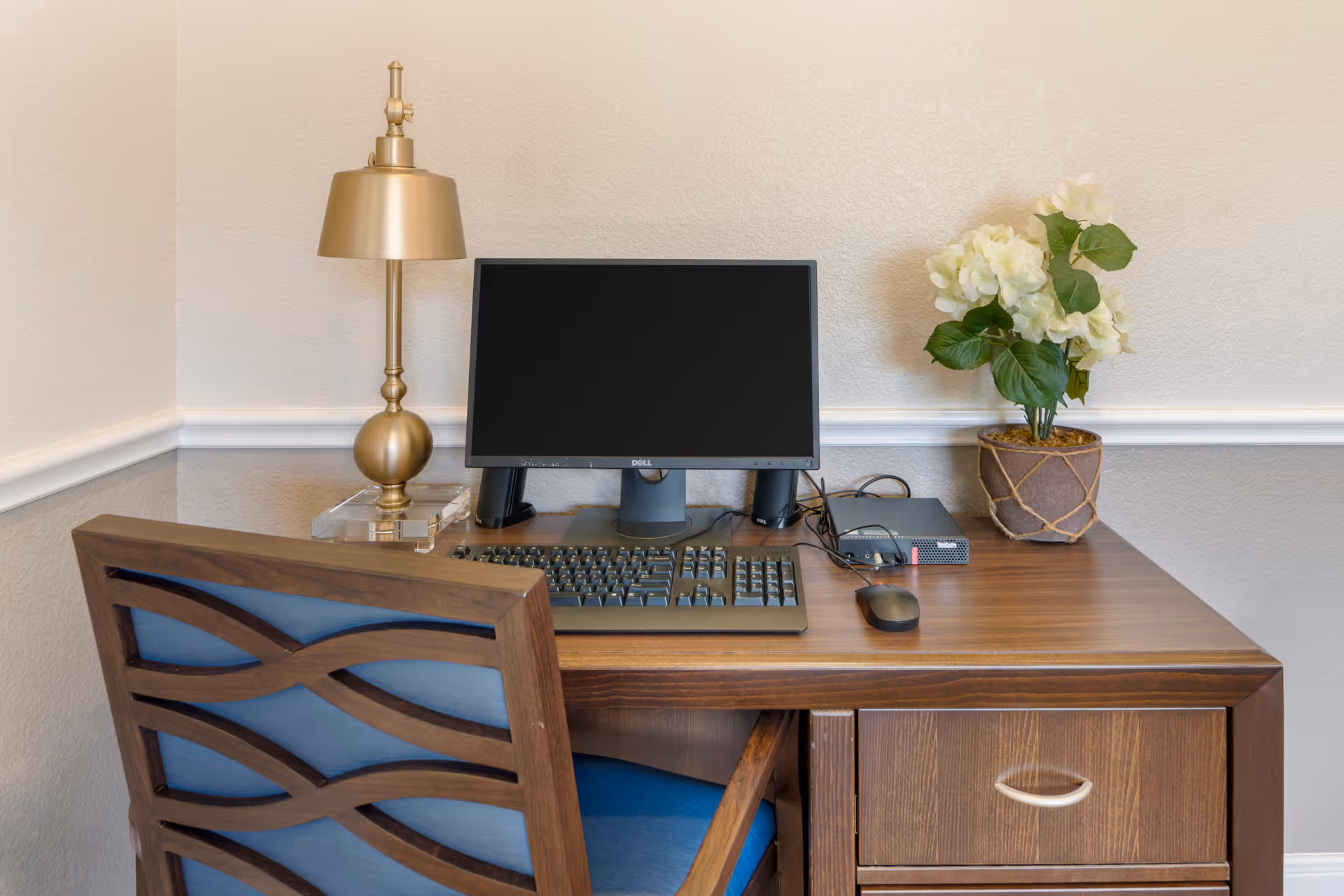 A wooden desk with a computer monitor, keyboard, mouse, a gold-colored lamp, and a potted plant with white flowers. A wooden chair with a blue cushion is positioned in front of the desk. The background features a beige textured wall with white trim.