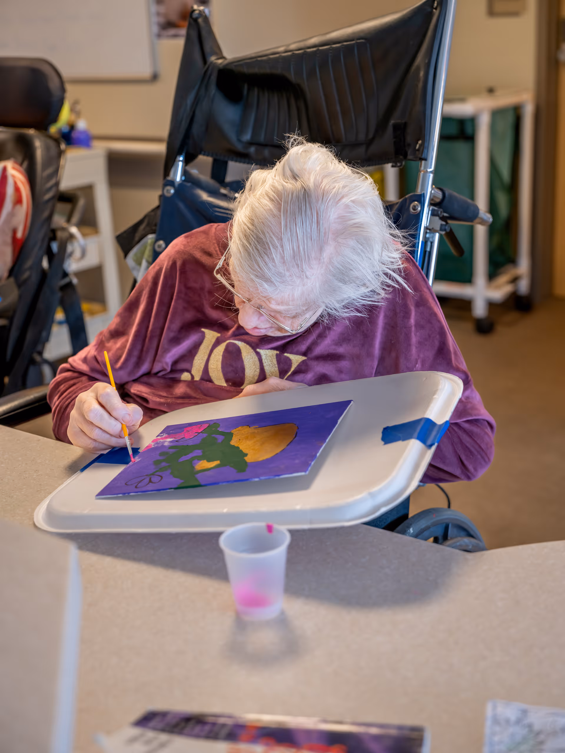 An elderly person in a wheelchair is painting a colorful picture on a tray table. The person is wearing glasses and a maroon shirt with the word 'JOY' printed on it. A small cup with pink paint is on the table in front of them.