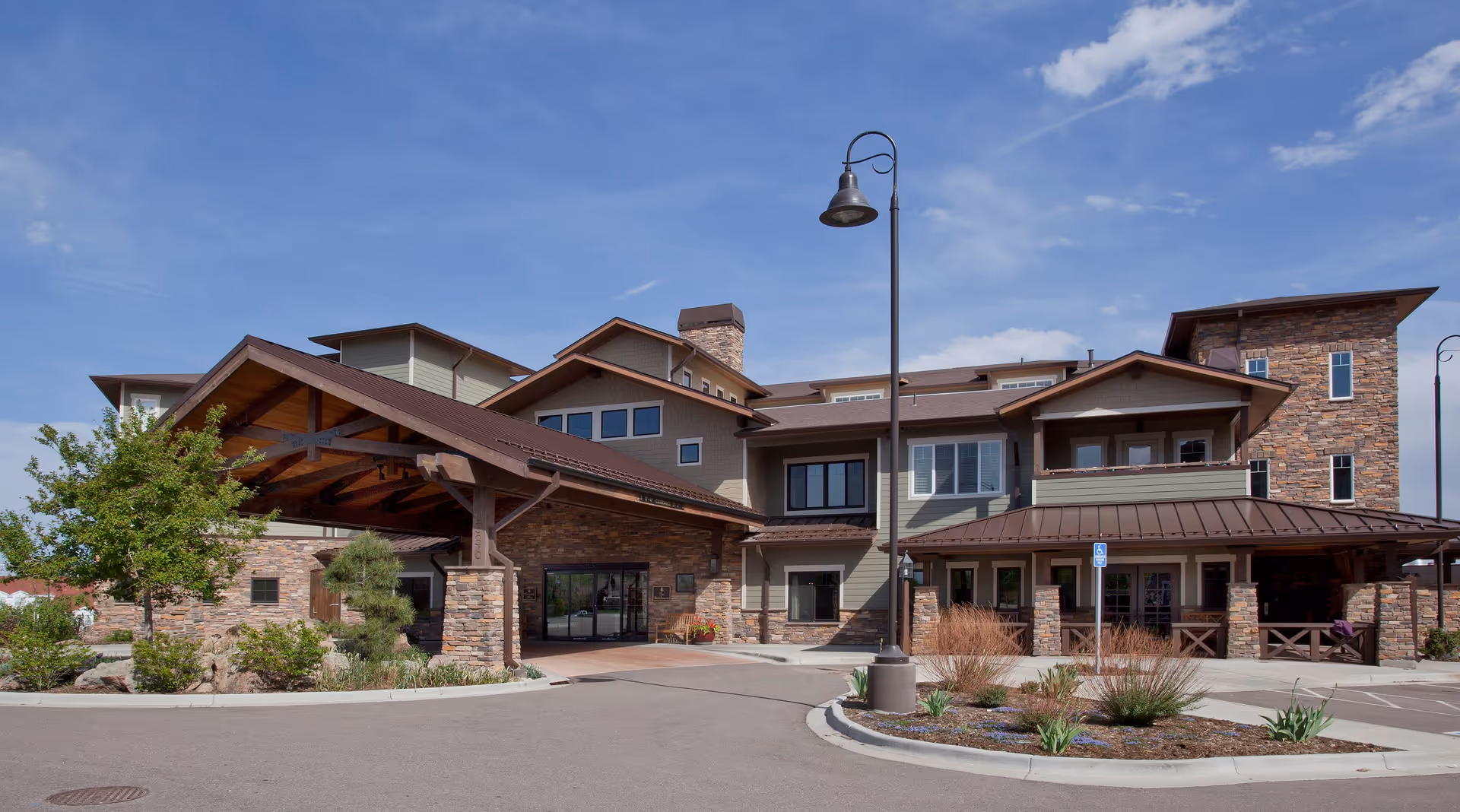 Exterior view of a senior living facility named The Residences at Balfour, featuring a large building with stone and wood accents, a covered entrance, landscaped greenery, and a clear blue sky.