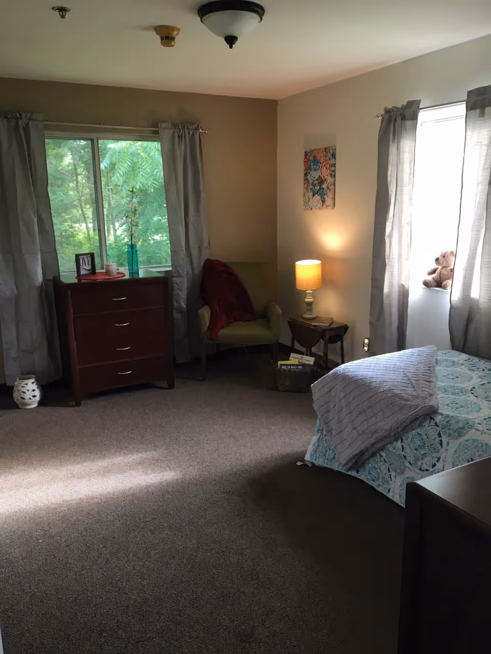 A cozy bedroom with a bed covered in a blue and white patterned bedspread and a gray blanket. There are two windows with gray curtains, one with a teddy bear on the windowsill. A wooden dresser with a vase and framed picture sits under one window. An armchair with a red throw blanket is next to a small side table with a lit lamp and books. The room has beige walls and carpeted floor.