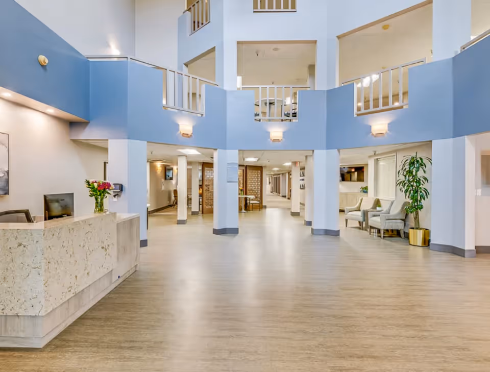 Spacious and well-lit lobby area of a senior living facility with a reception desk on the left, decorated with a vase of flowers. The lobby features light wood flooring, blue and white walls, seating area with chairs and a potted plant on the right, and an open second-floor balcony overlooking the space.