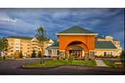 Exterior view of Longwood At Oakmont Retirement Community showing a large building with a covered entrance, green roof, and multiple windows under a cloudy sky. There is a paved driveway and landscaped greenery in front.