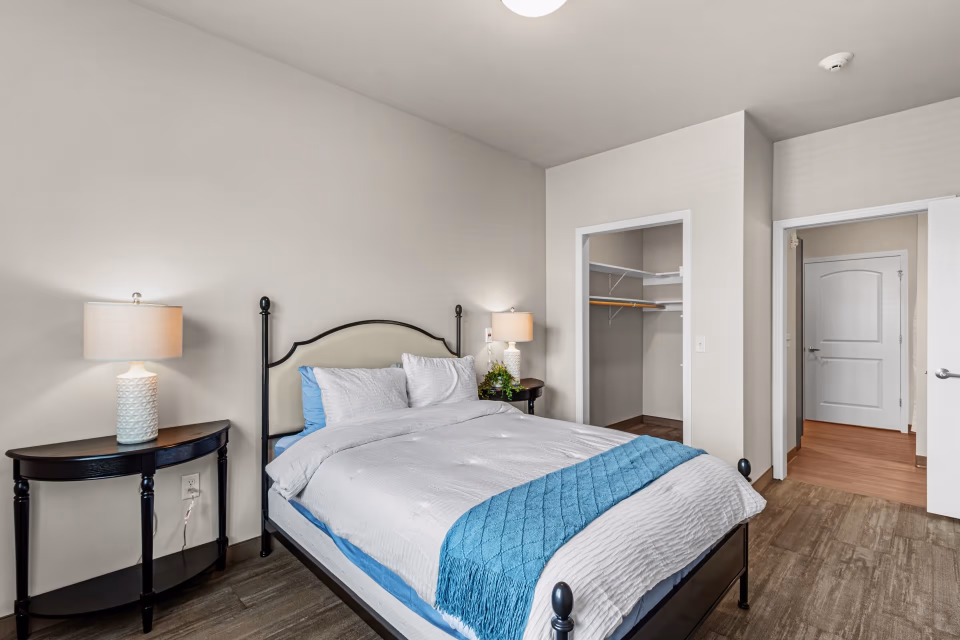 A neatly made bed with white and blue bedding in a bedroom. There are two bedside tables with lamps on each side of the bed. The room has light-colored walls and wood flooring. An open closet and a doorway leading to another room are visible.