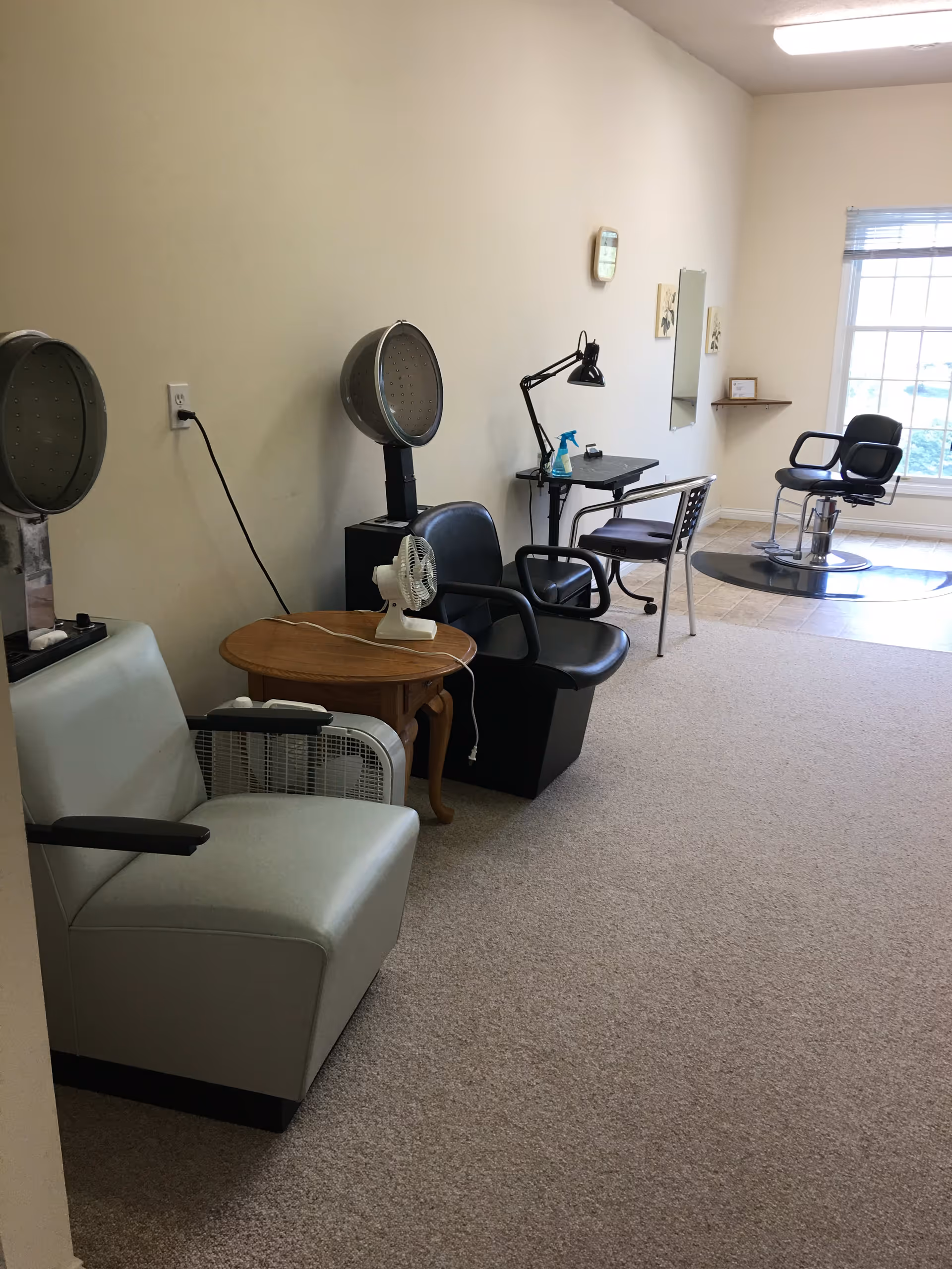 Interior view of a salon area with hair drying chairs, a small wooden table with a fan, a desk with a lamp and chair, and a styling chair near a large window with blinds.