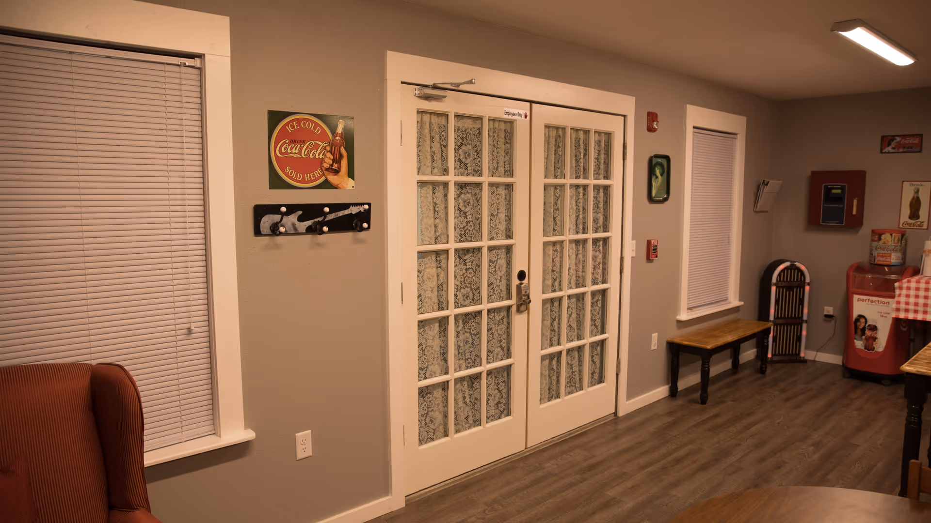 Interior view of a room with double French doors featuring lace curtains. The room has gray walls, wood flooring, and two windows with closed blinds. There is a red armchair on the left, a wooden bench under one window, a vintage-style Coca-Cola sign on the wall, a coat rack, a fire alarm, and a red vending machine in the corner.