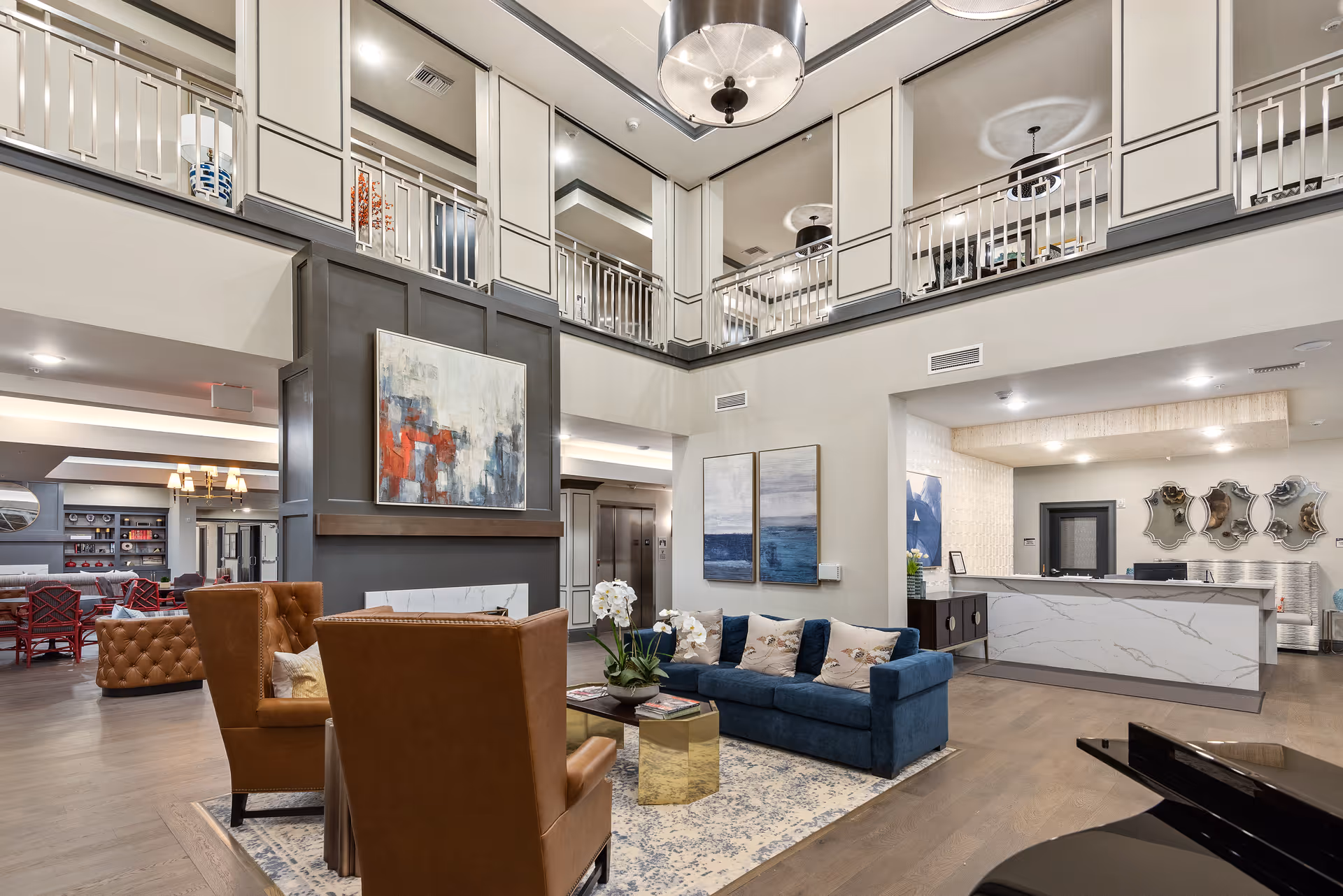 Spacious and modern senior living facility common area with high ceilings and a mezzanine level. The room features a blue sofa with decorative pillows, two brown leather armchairs, a coffee table with a plant, and a large abstract painting above a fireplace. In the background, there is a reception desk with three decorative mirrors on the wall and additional seating areas with red chairs and a chandelier.