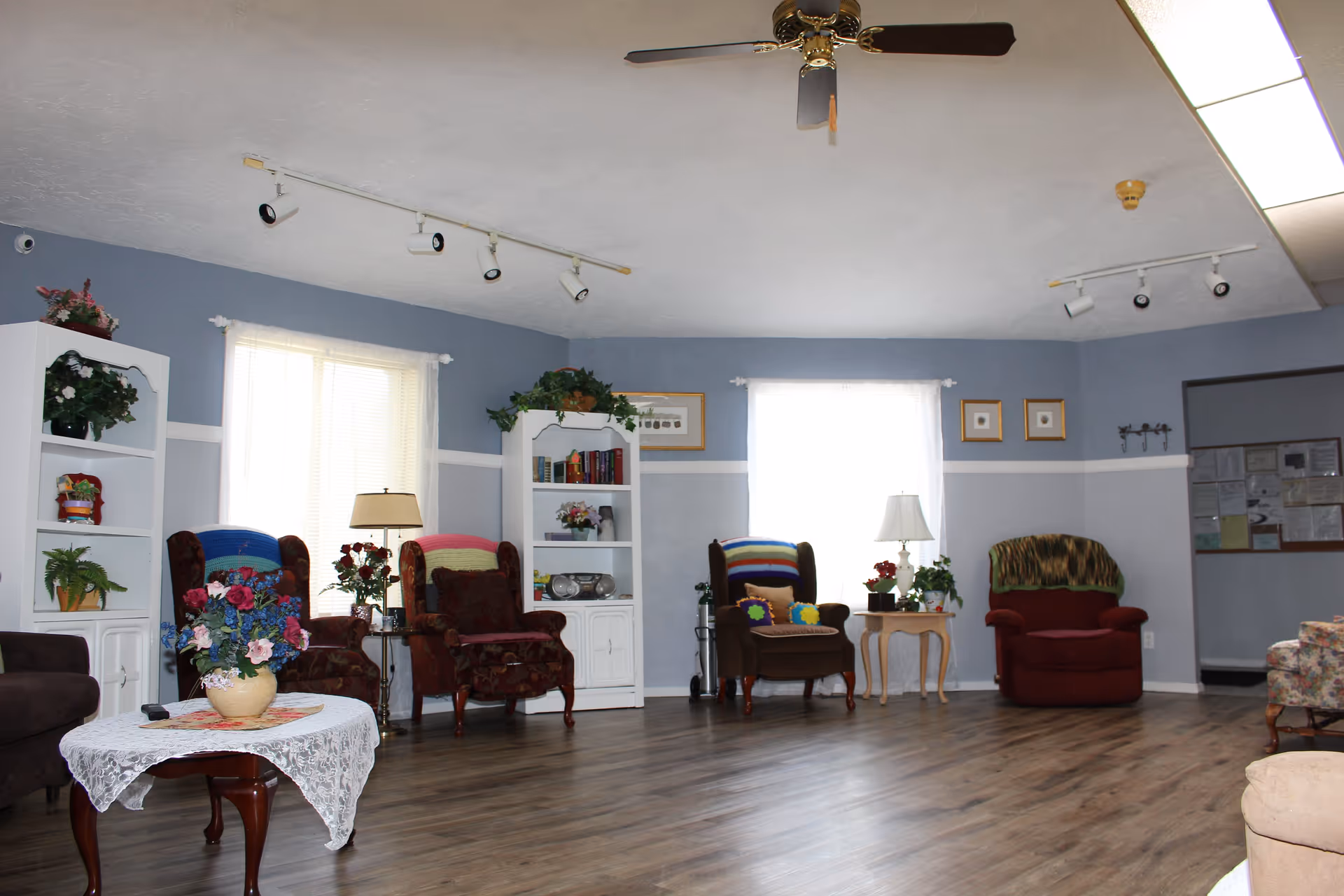 A spacious living room with light blue walls and wood flooring. The room features several armchairs with colorful blankets and cushions, two white bookshelves with plants and books, a coffee table with a lace tablecloth and a flower arrangement, and two windows with sheer white curtains. Ceiling lights and a ceiling fan are visible above.