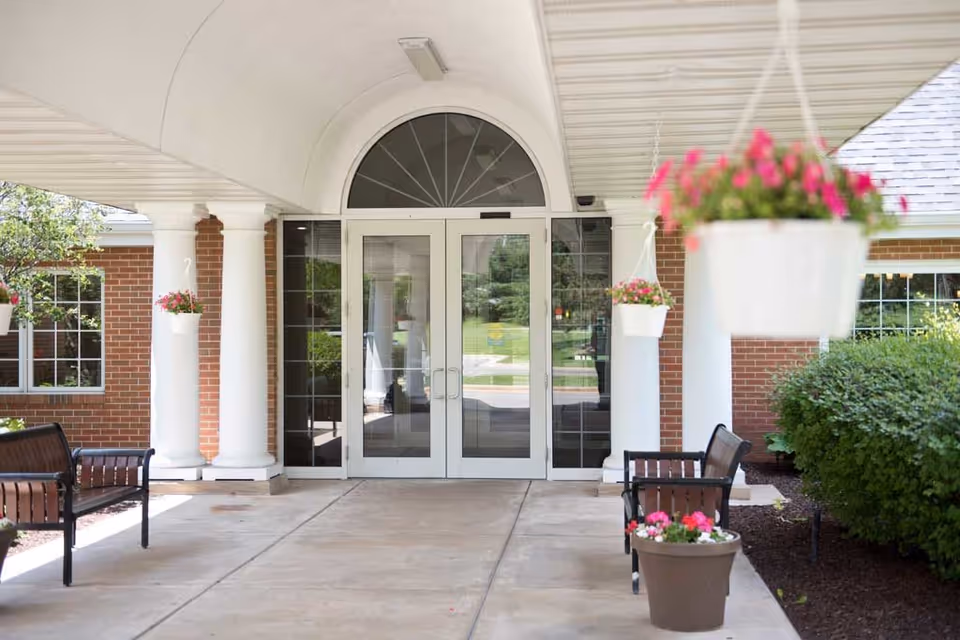 Covered entrance with double glass doors flanked by white columns, benches, and hanging flower baskets.