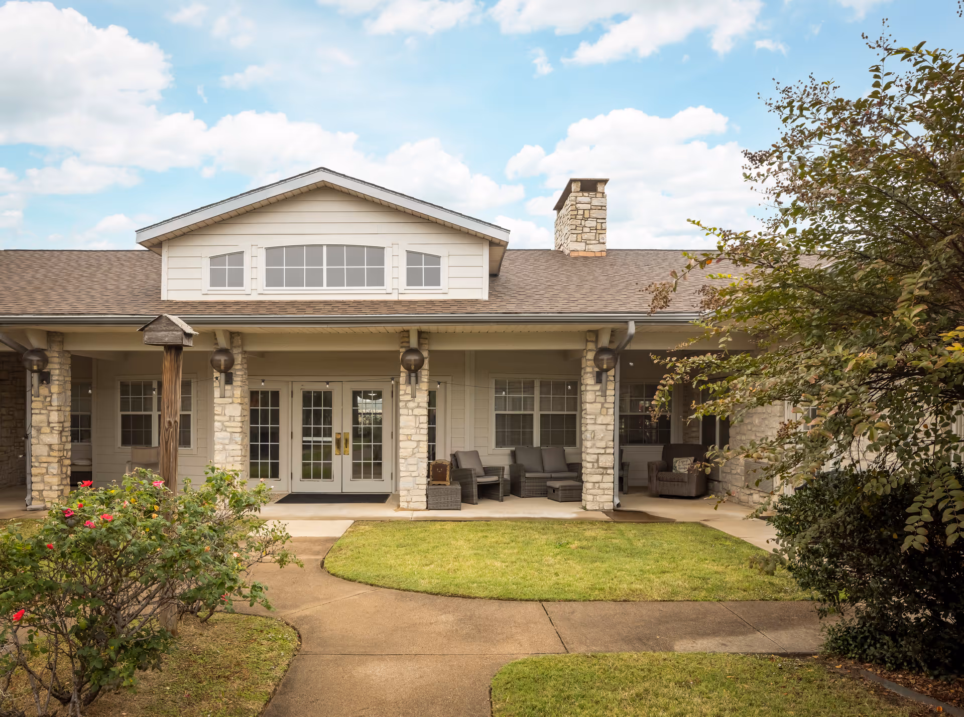 Front exterior view of a single-story building with stone pillars, a covered porch with outdoor seating, and a well-maintained lawn and garden under a partly cloudy sky.