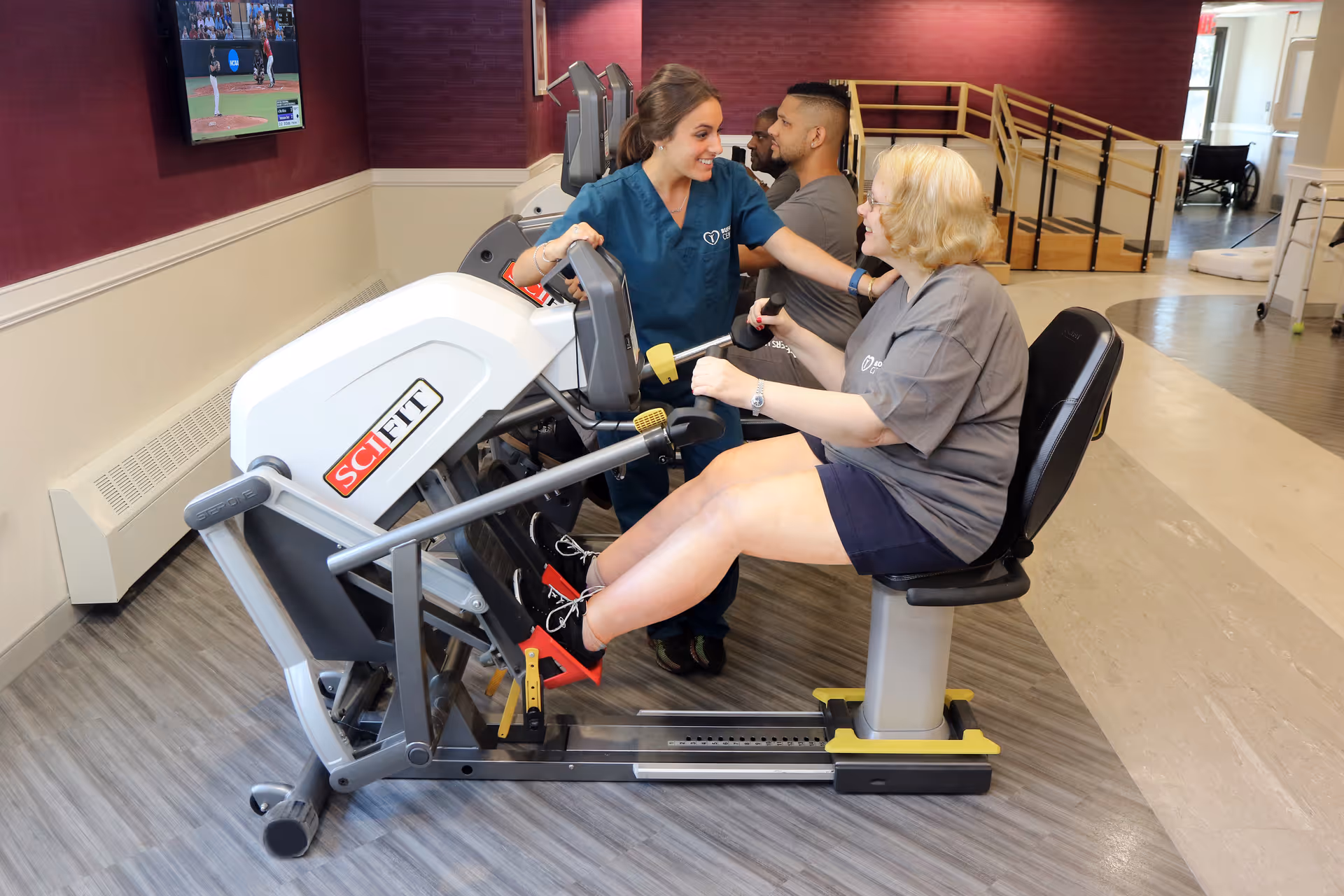 A woman in a gray shirt is using a SciFit exercise machine in a rehabilitation facility while a staff member in blue scrubs encourages her. Another man is using a similar machine in the background. A TV is mounted on the wall showing a baseball game.