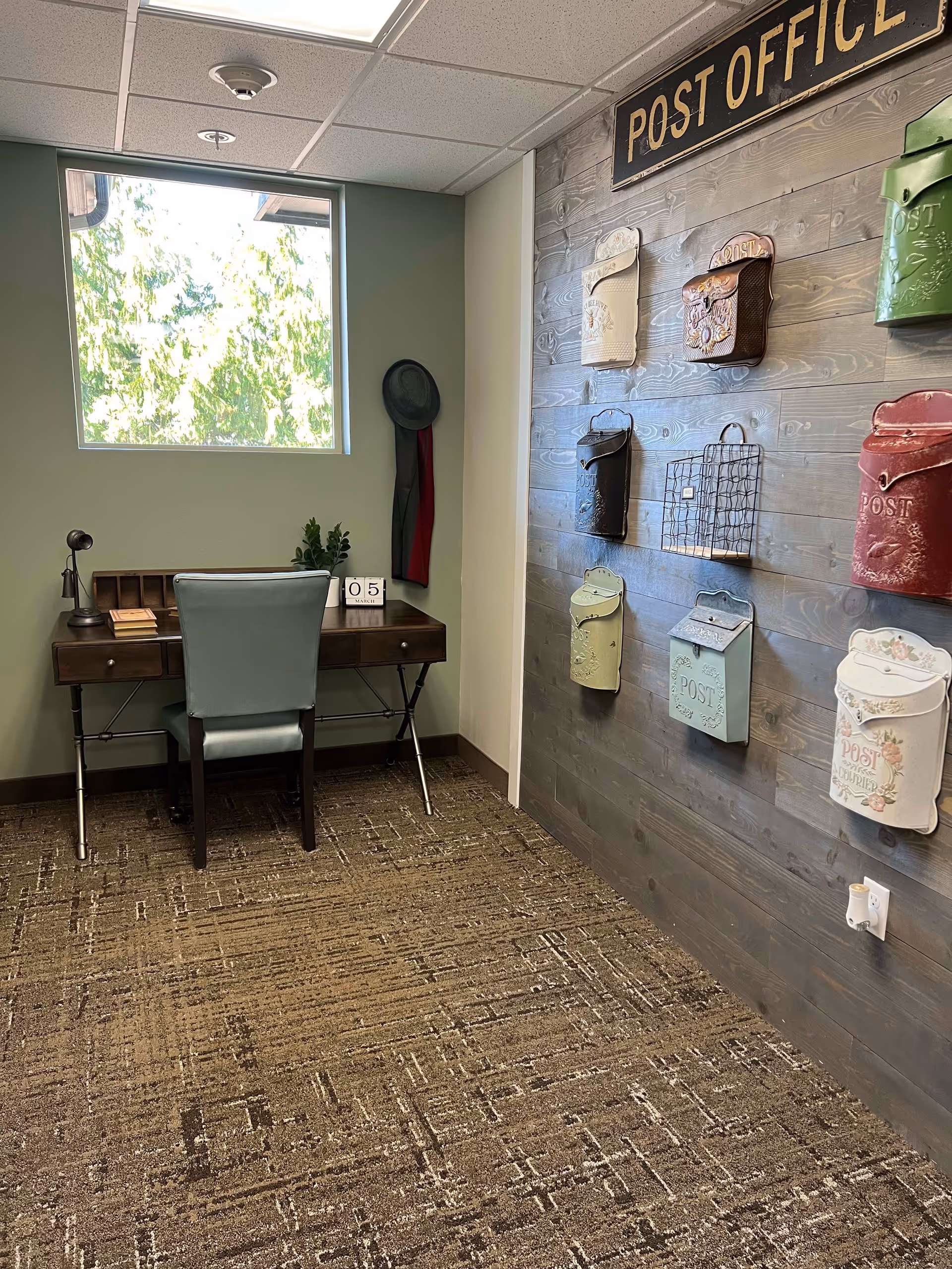 A small room with a green wall and a window letting in natural light. There is a wooden desk with a green chair, a small plant, a desk calendar showing the number 05, and a few books. On the adjacent wall, there is a decorative display of various vintage-style mailboxes mounted on wooden paneling under a sign that reads 'POST OFFICE'.