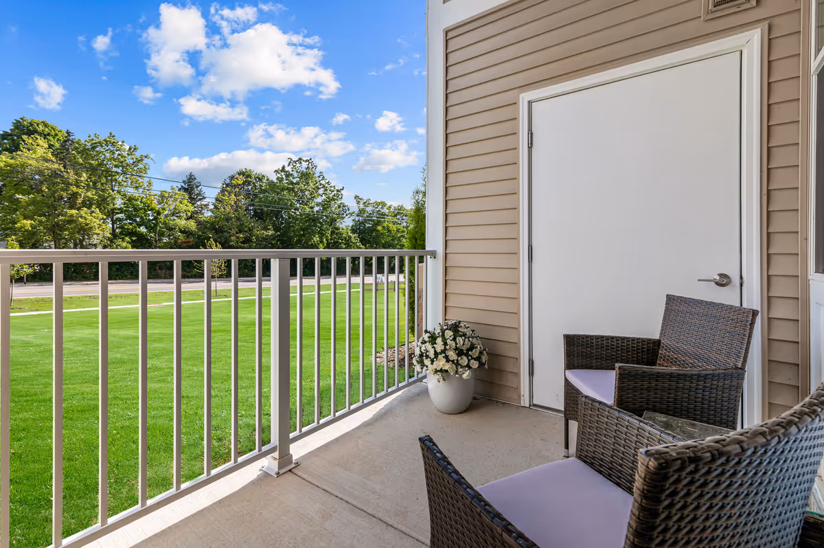 A small outdoor balcony area with two wicker chairs with light purple cushions, a white potted plant with white flowers, beige siding on the building, a white door, and a white railing overlooking a green lawn and trees under a blue sky with scattered clouds.